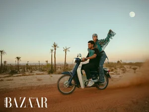 A man driving a vintage blue moped on a desert dirt road with fashion model Tilila Oulhaj seated behind in a patterned headscarf.