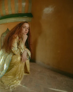 Model with long red hair crouching in a yellow patterned garment inside a warmly lit traditional interior space