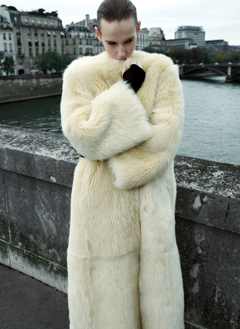 Fashionable adult woman in a long, ivory-colored faux fur coat standing on a bridge overlooking the Seine River in Paris.