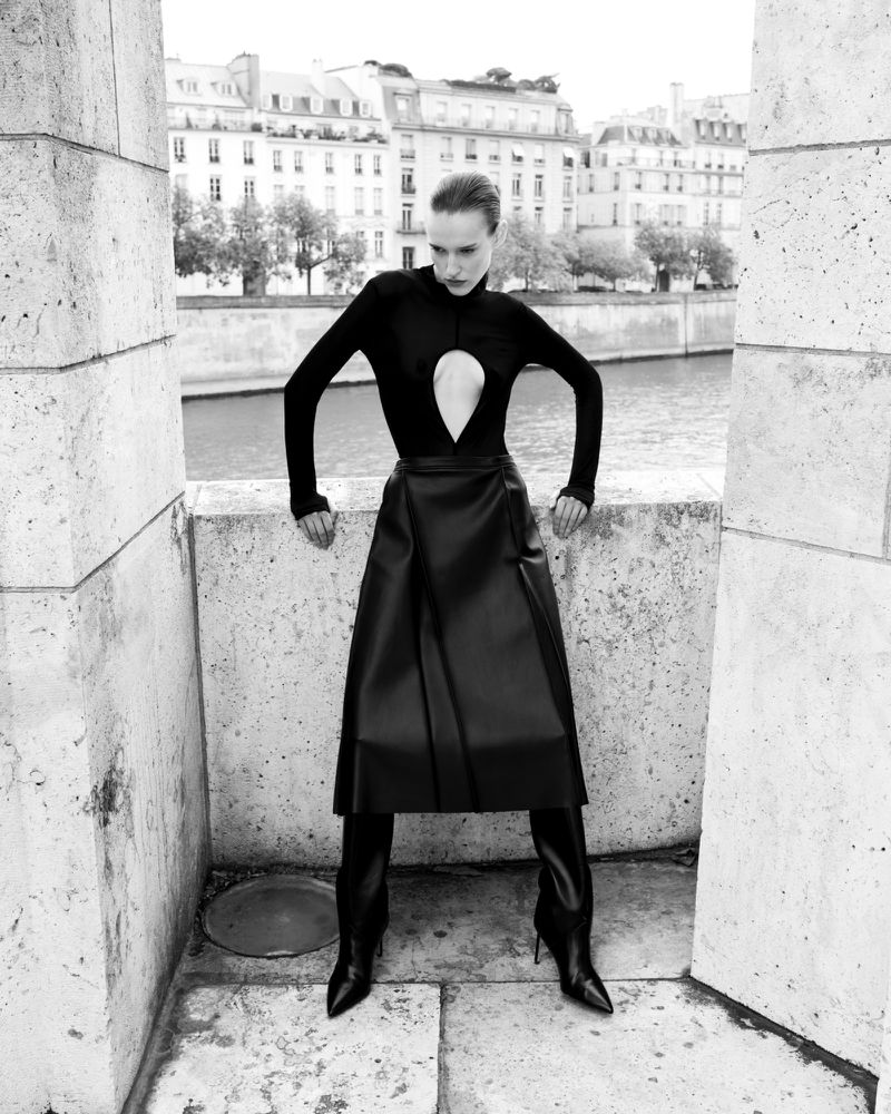Monochrome portrait of a model wearing a black top and leather skirt standing on a stone bridge with buildings in the background.