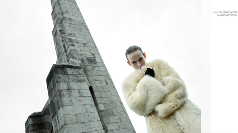 Woman in white fur coat poses with geometric stone monument under bright sky. Low-angle shot emphasizes monument's height.