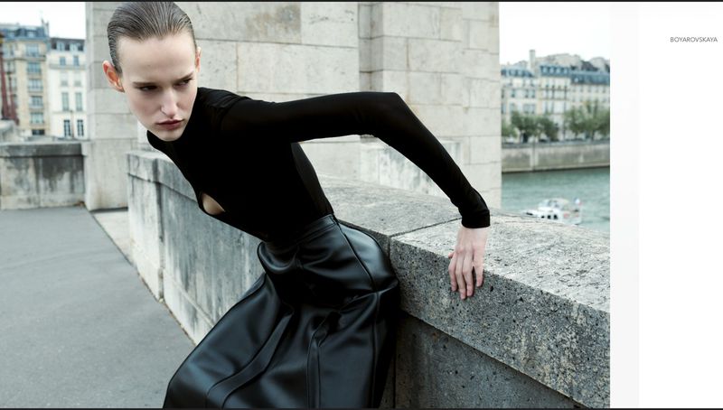 Model in black top and leather skirt leaning on a stone bridge overlooking a river