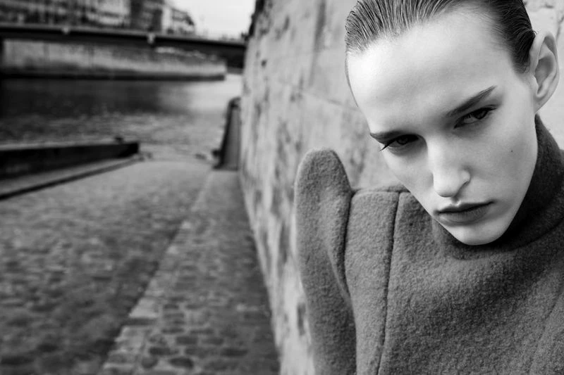 Black and white portrait of a slender woman with slicked-back hair, wearing a gray turtleneck coat, posing outdoors by a stone wall and river.