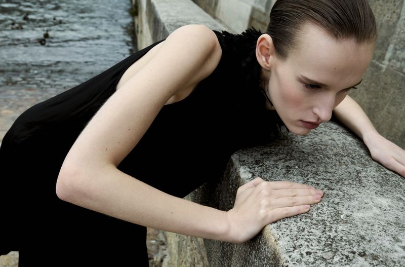 Fashion shot of a woman in a black dress leaning on a stone wall by a river, with wet hair and neutral expression, and dark, moody lighting.
