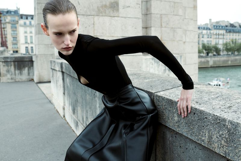 Fashion editorial shot of a woman in a black top and black skirt leaning against a stone wall with a cityscape in the background.