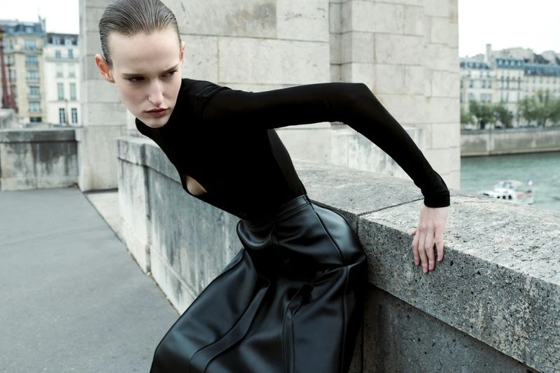 Fashion editorial shot of a woman in a black top and black skirt leaning against a stone wall with a cityscape in the background.