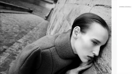 Black and white profile portrait of model Alex Shibaeva resting her head against a stone wall in a high-collar coat.