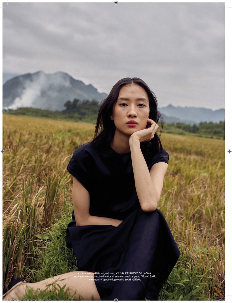 Asian woman in black dress poses in golden rice field under cloudy sky with mountains in the distance. Editorial fashion shot.