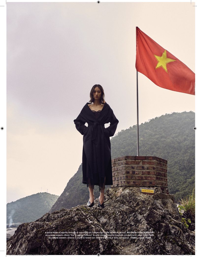 A female model in a black dress stands on a rocky outcrop with a red and yellow Vietnamese flag waving in the background.