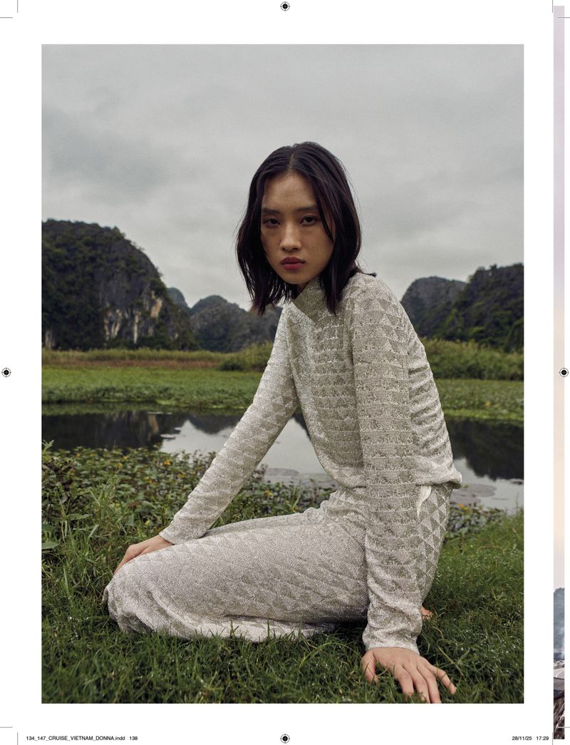 A young woman in a textured, long-sleeve top and skirt poses in a grassy field with a lake and karst mountains in the background.