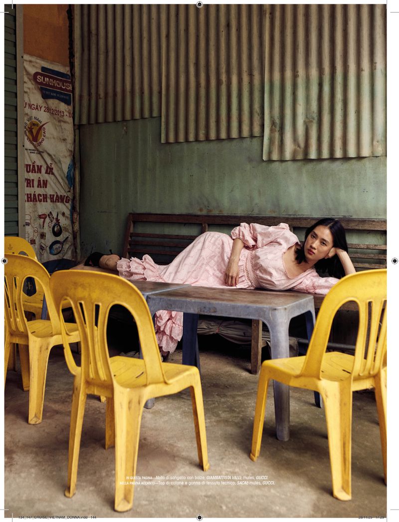 A woman in a long-sleeved pink dress poses on a table surrounded by yellow plastic chairs, in an outdoor, vintage-style setting.