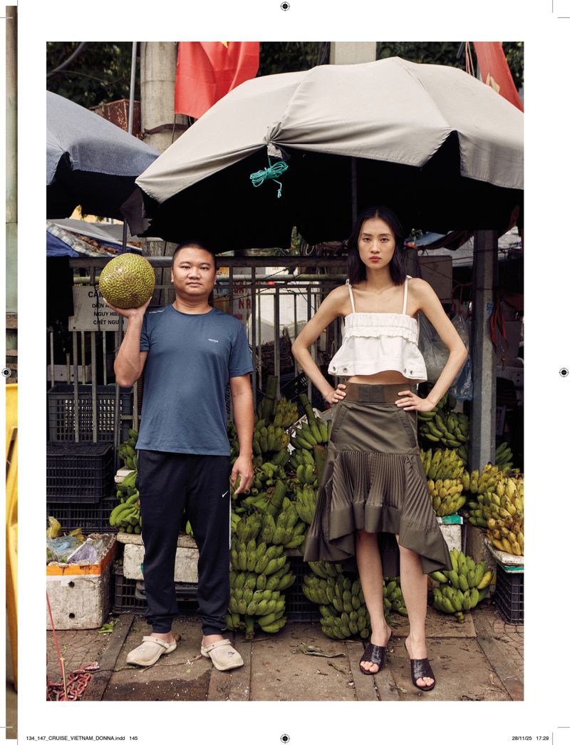 Two people posing in front of a fruit stand with bananas and other produce, beneath an umbrella in an outdoor market setting.