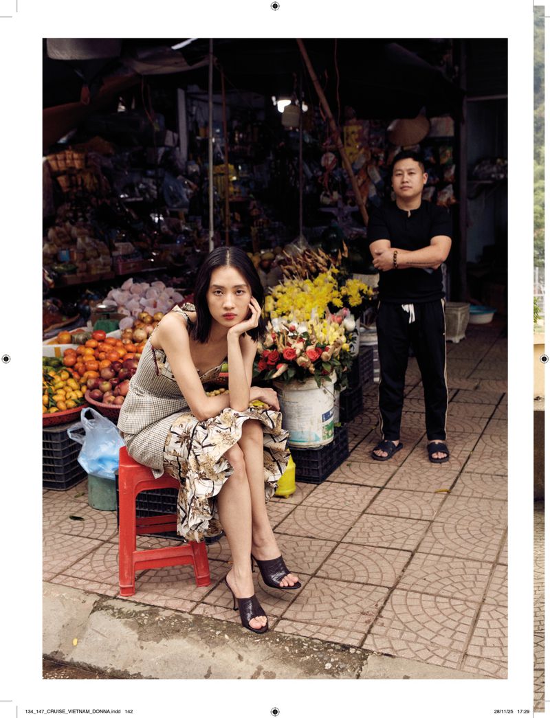 Asian woman in a dress sits on a red stool near flowers and produce at an outdoor market, with a man standing in the background.
