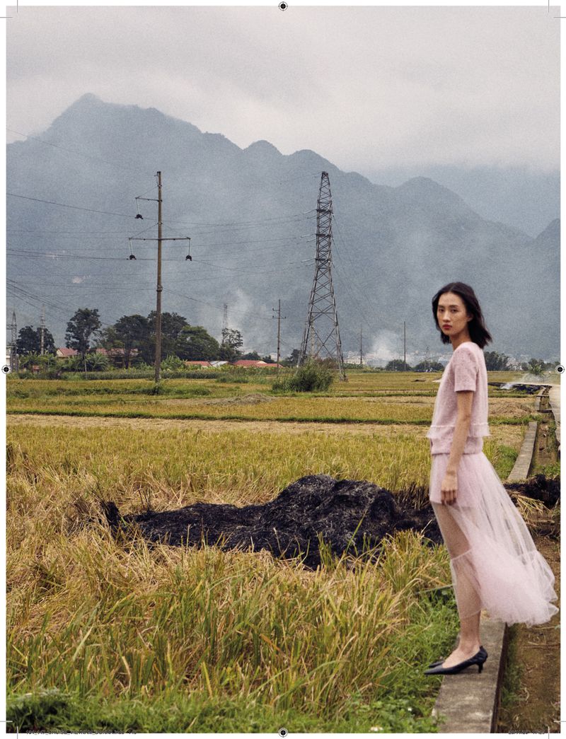A young woman stands on a path, posing in a soft pink dress against a scenic backdrop of rice fields and distant mountains.