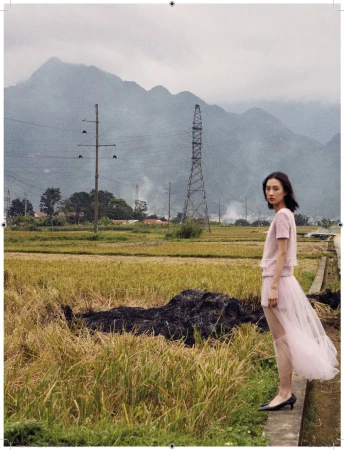 A young woman stands on a path, posing in a soft pink dress against a scenic backdrop of rice fields and distant mountains.