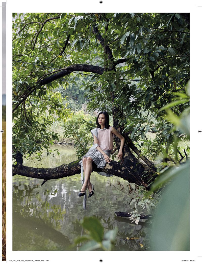Woman in a pink top and gray pleated skirt sits on a tree branch over a lake surrounded by green foliage in an outdoor setting.