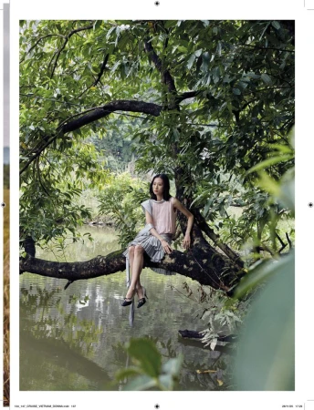 Woman in a pink top and gray pleated skirt sits on a tree branch over a lake surrounded by green foliage in an outdoor setting.