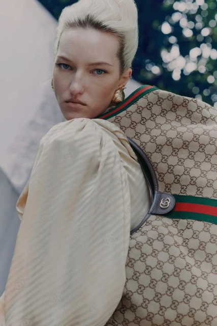 Blonde model wearing Gucci satchel bag and light peach dress with sleeves, posing against a blurred outdoor setting, fashionable.