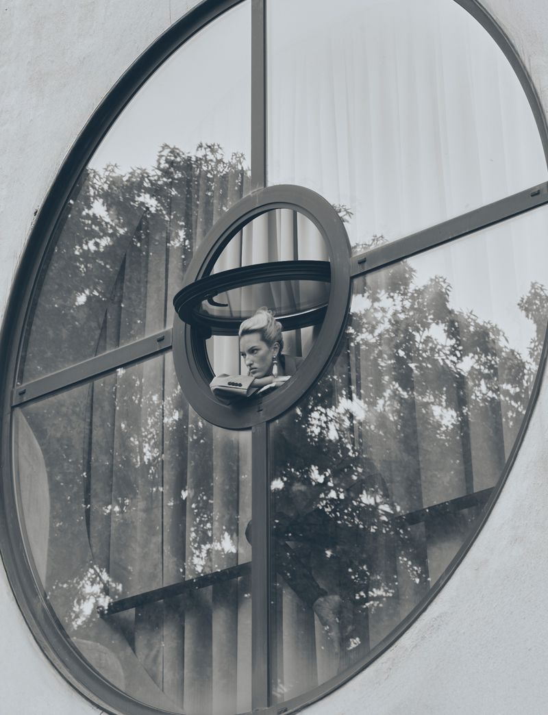 Monochrome portrait of a woman with short hair, wearing earrings, and reading, framed by an architectural round window and trees.