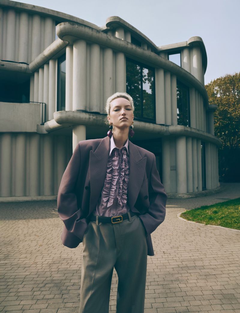 A woman poses in a lilac suit outside an architectural building on a brick surface during the daytime. Outdoor fashion editorial.