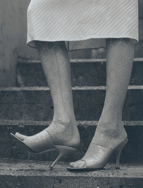 Close-up of woman's legs and feet on steps, wearing patterned hosiery and heeled sandals, in monochromatic grey-scale.