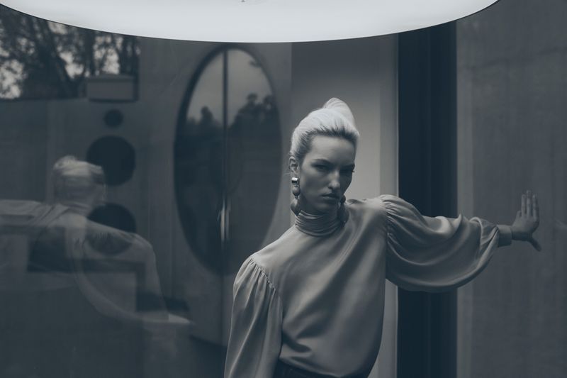 Monochrome portrait of a blonde woman in a satin top with statement earrings, posing indoors with minimalist decor and geometric elements.