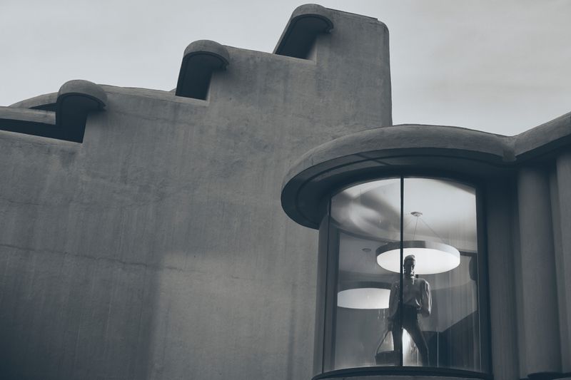 Monochrome architectural shot of a mannequin in a glass-enclosed space with concrete building and round lights in the background.
