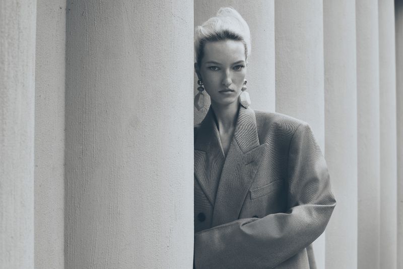 Portrait of a blonde woman in an oversized blazer with a serious expression, posing next to fluted white pillars in soft, diffused lighting.