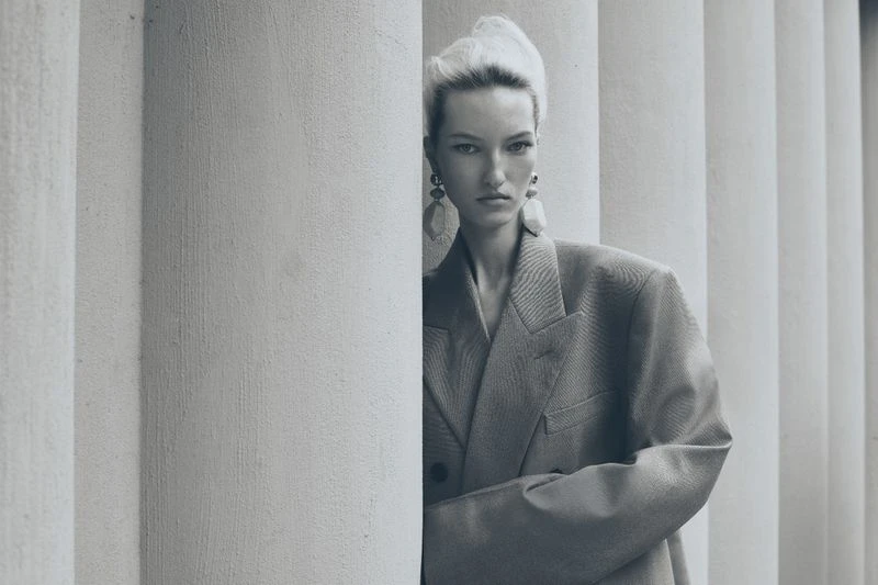 Portrait of a blonde woman in an oversized blazer with a serious expression, posing next to fluted white pillars in soft, diffused lighting.