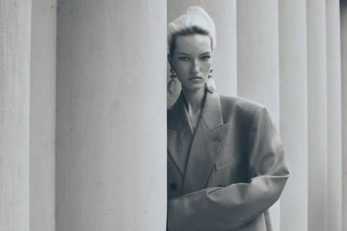 Portrait of a blonde woman in an oversized blazer with a serious expression, posing next to fluted white pillars in soft, diffused lighting.