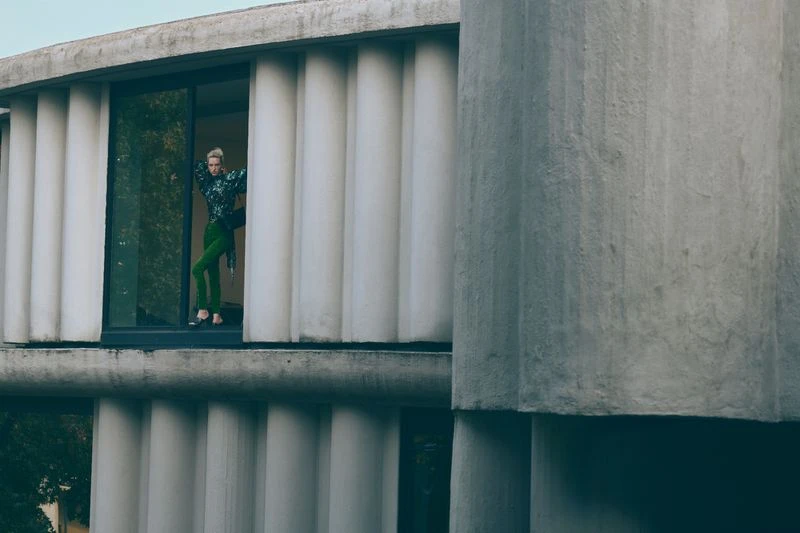 A woman in green pants and a floral shirt is standing in an opening in a modern concrete building, posing for a fashion photograph.