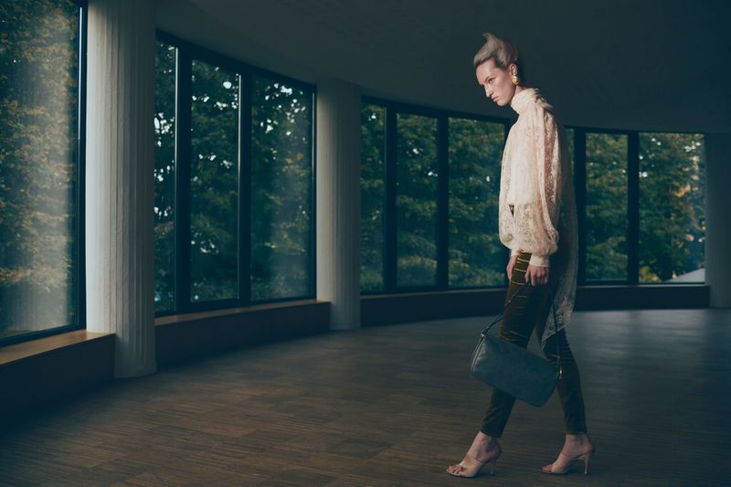 Fashion photo of a stylish woman in a lace blouse and green trousers walking in a modern building with large windows and hardwood floors.