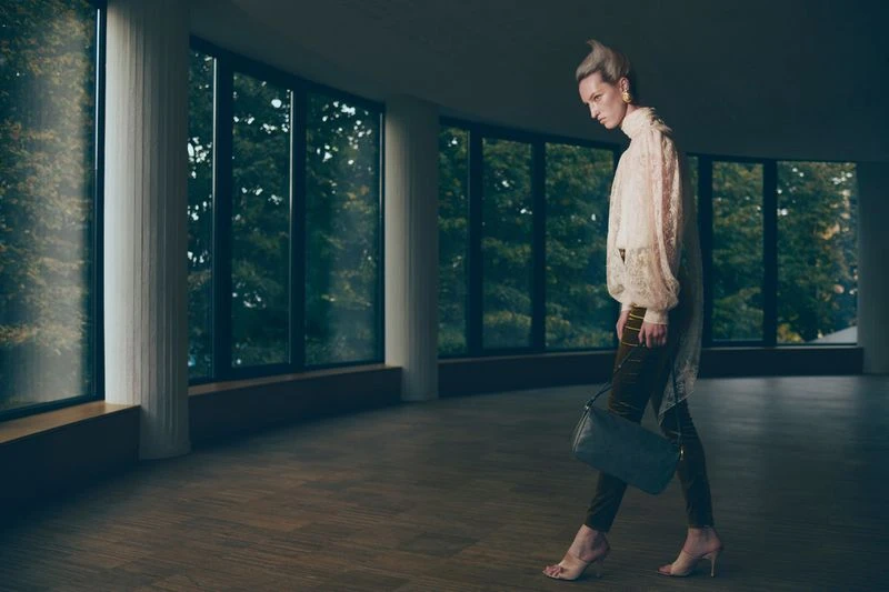 Fashion photo of a stylish woman in a lace blouse and green trousers walking in a modern building with large windows and hardwood floors.