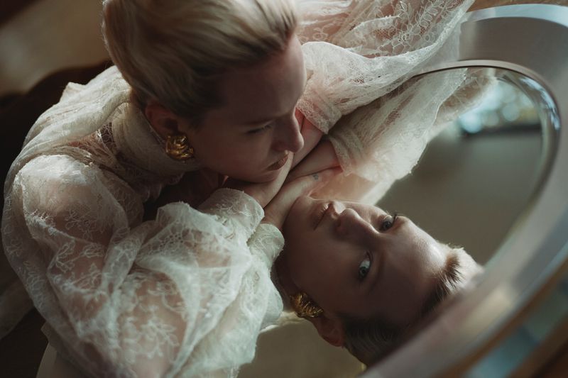 An adult female poses indoors in front of a mirror wearing a white lace dress and large gold earrings in a medium shot.
