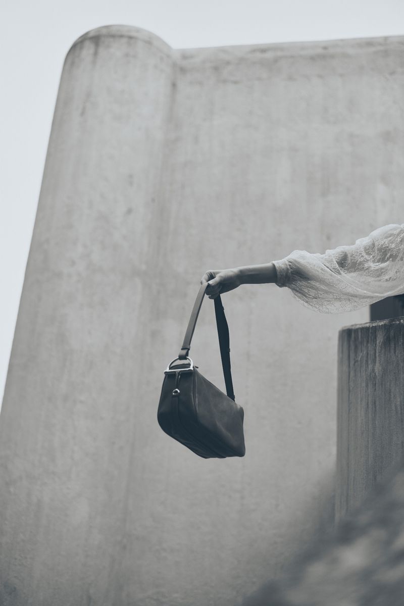 Black leather bag being held by a person with long sleeves against the backdrop of a concrete structure on an overcast day.
