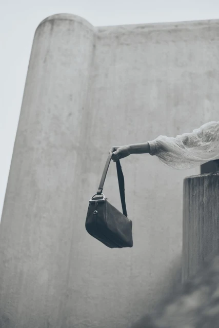 Black leather bag being held by a person with long sleeves against the backdrop of a concrete structure on an overcast day.