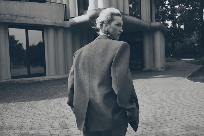 Monochrome portrait of a woman in a blazer standing on a brick sidewalk in front of an architectural building with green trees nearby.