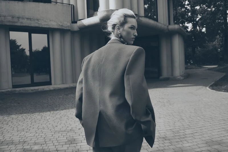 Monochrome portrait of a woman in a blazer standing on a brick sidewalk in front of an architectural building with green trees nearby.