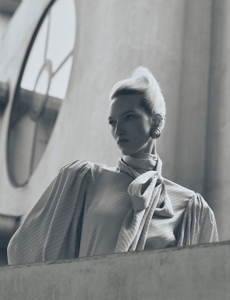 Monochrome portrait of a blonde woman in a vintage-style satin blouse with a large bow, posing indoors with a large arched window.