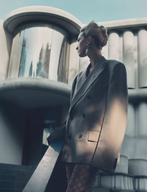 Model poses outdoors in oversized grey blazer and patterned tights in front of concrete building on an overcast day, looking away.