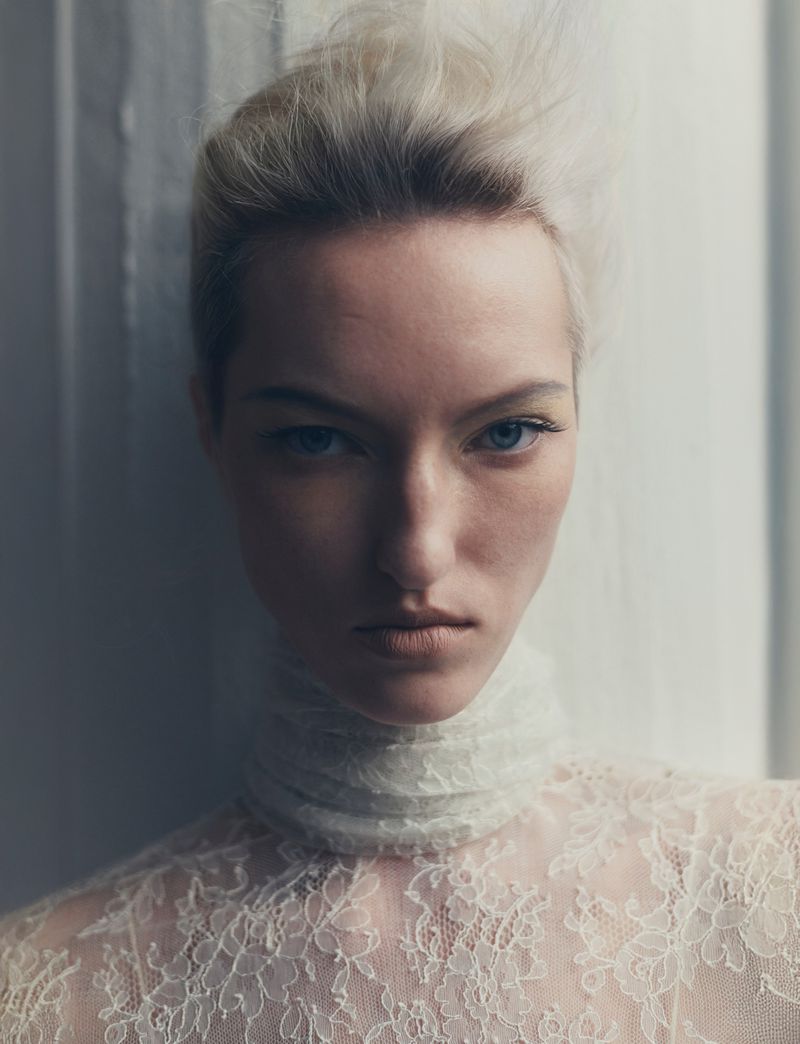 Close-up portrait of a light-haired adult woman with blue eyes and fair skin, wearing a white lace top and standing indoors.