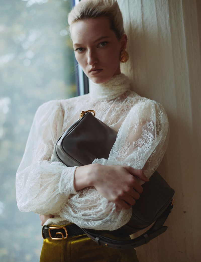 Indoor shot of woman with blonde hair, wearing a lace top with yellow-green pants, and holding a dark gray leather handbag.