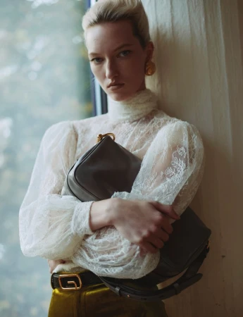 Indoor shot of woman with blonde hair, wearing a lace top with yellow-green pants, and holding a dark gray leather handbag.