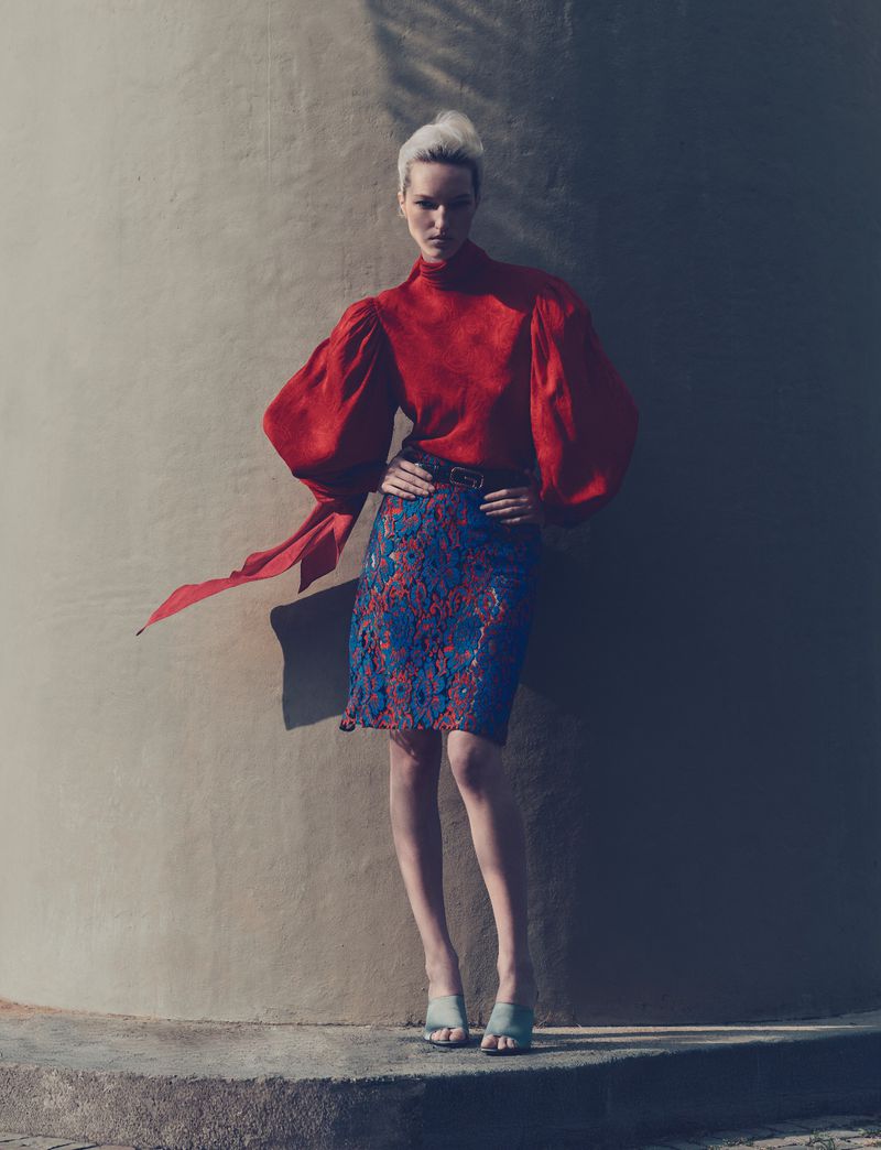 Woman in a red top and floral skirt posing against a wall with shadows, fashion editorial shot with a neutral color palette.