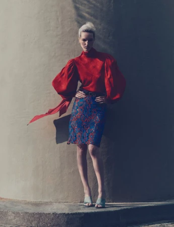 Woman in a red top and floral skirt posing against a wall with shadows, fashion editorial shot with a neutral color palette.