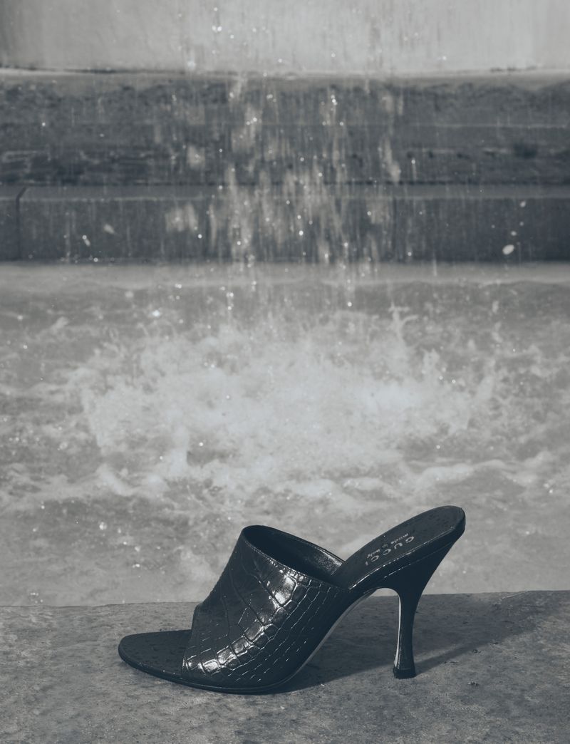 Black textured heeled sandal positioned near a stone fountain with running water in a monochromatic setting, fashion editorial.
