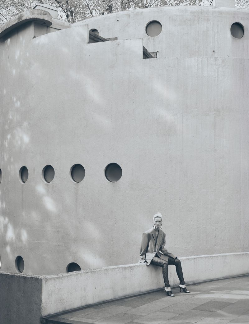 A stylish woman poses seated on a wall, wearing a blazer and skinny jeans, outside a modern brutalist building with round windows.