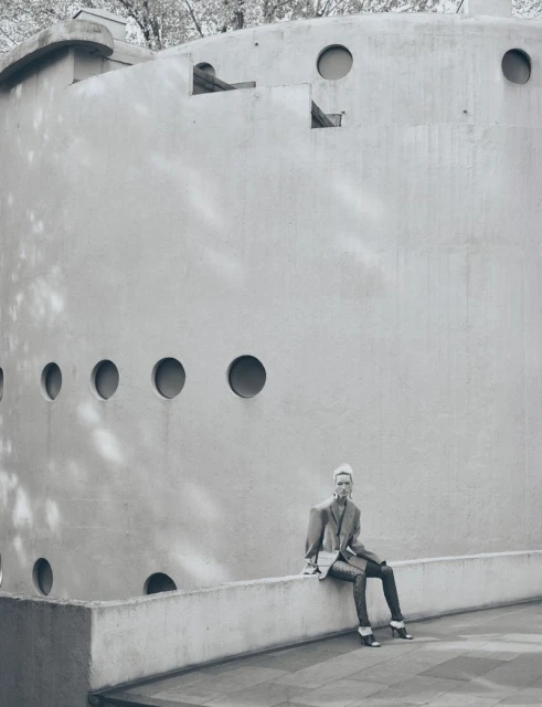 A stylish woman poses seated on a wall, wearing a blazer and skinny jeans, outside a modern brutalist building with round windows.