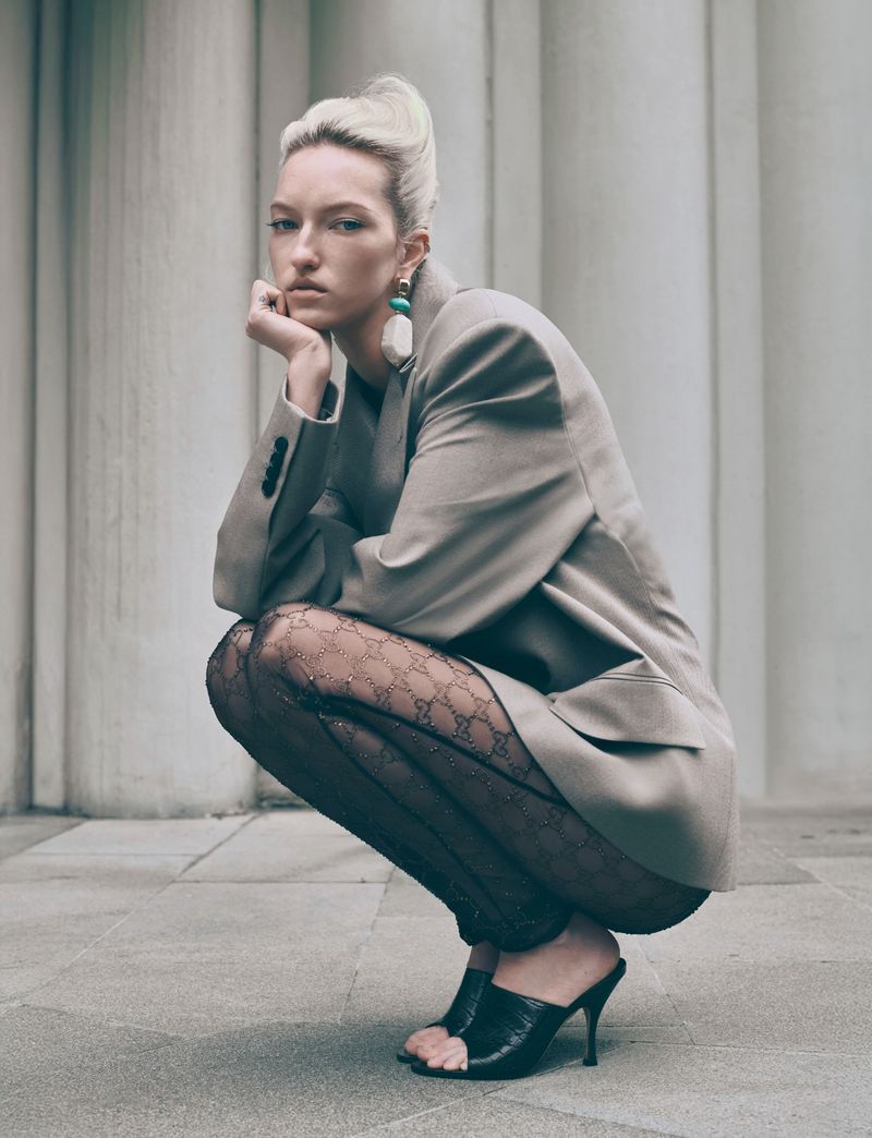 Fashion shot of a blond woman in an oversized blazer, patterned tights, and heeled mules posing outdoors in front of a column backdrop.