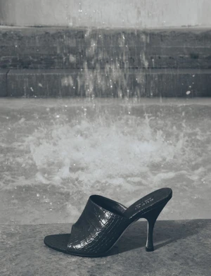 Black crocodile-embossed high-heeled mule resting on a stone ledge in front of splashing fountain water.